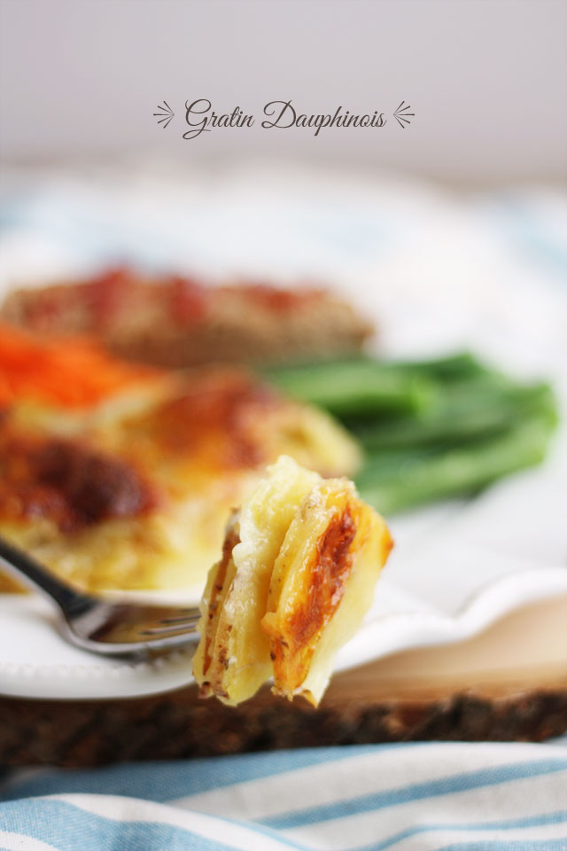 sliced potatoes baked with a creamy, cheesy sauce, with other foods in the background on a plate