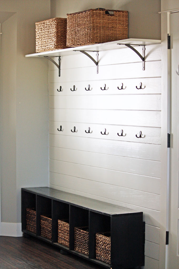 A mudroom featuring a white plank wall with two rows of double coat hooks, an upper shelf with oil-rubbed bronze brackets, and a bench with storage baskets | modern farmhouse inspiration